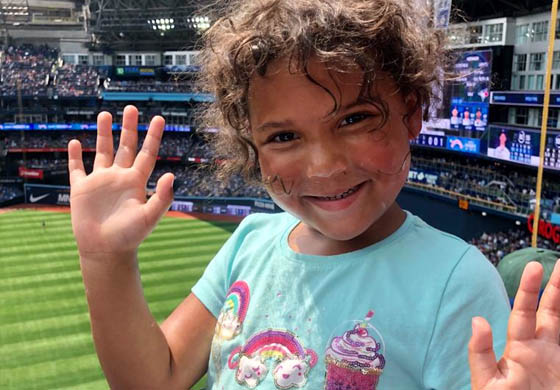 Little girl in a blue shirt smiling with her hands up at a Blue Jays game in Toronto