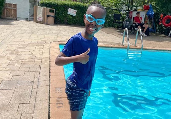 Young black boy smiling and with his thumb up, standing on the edge of a pool, dripping wet